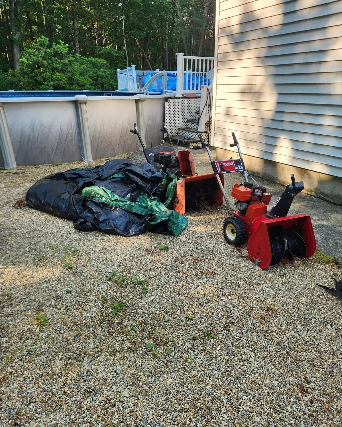 Two old snowblowers and tarps in a gravel yard beside a pool